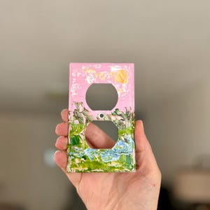 May include: A rectangular light switch wall plate featuring a painted landscape design. The top section is light pink with a small yellow sun, while the bottom section displays green, brown, and blue textured paint strokes representing a landscape.