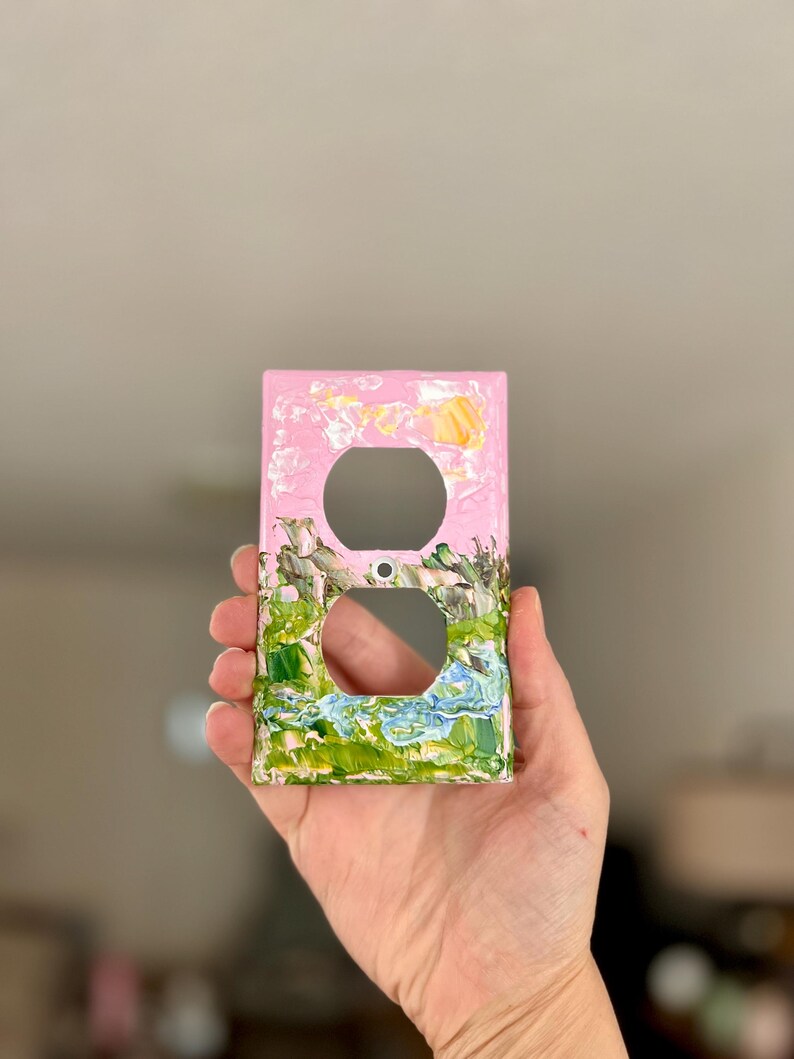 May include: A rectangular light switch wall plate featuring a painted landscape design. The top section is light pink with a small yellow sun, while the bottom section displays green, brown, and blue textured paint strokes representing a landscape.