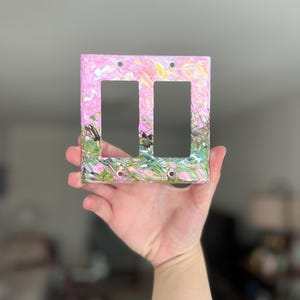 May include: A rectangular double light switch cover plate featuring a textured, impasto-style paint application. The surface displays a blend of pastel pink, soft yellow, and vibrant green hues, with visible brushstrokes creating a layered, abstract effect.