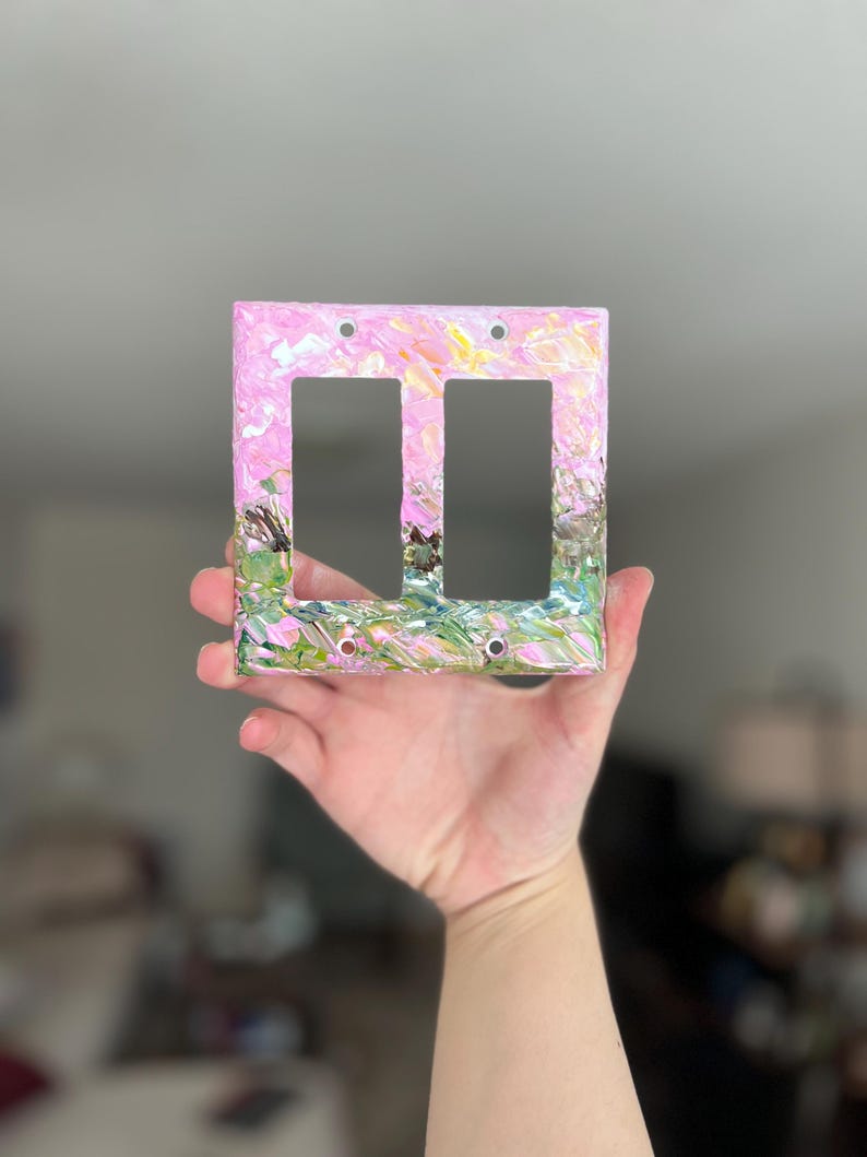 May include: A rectangular double light switch cover plate featuring a textured, impasto-style paint application. The surface displays a blend of pastel pink, soft yellow, and vibrant green hues, with visible brushstrokes creating a layered, abstract effect.
