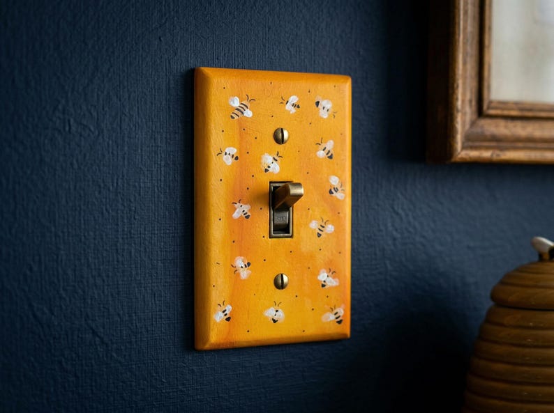 Puede incluir: Una placa de interruptor de luz rectangular con un fondo degradado en tonos amarillo y naranja c&aacute;lidos, que presenta un patr&oacute;n repetitivo de peque&ntilde;as abejas pintadas a mano en blanco y negro. Incluye un interruptor central y dos tornillos color lat&oacute;n.