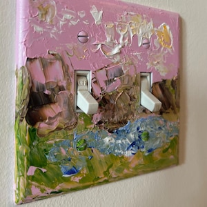 May include: A double light switch plate featuring a thick, textured impasto oil painting application. The design displays a landscape with pink sky, brown rock formations, and green grass, accented by white and blue paint strokes and two white toggles.