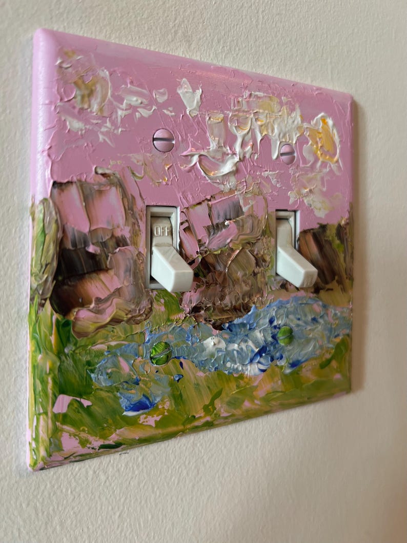 May include: A double light switch plate featuring a thick, textured impasto oil painting application. The design displays a landscape with pink sky, brown rock formations, and green grass, accented by white and blue paint strokes and two white toggles.