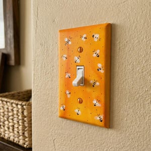 Puede incluir: Una placa de interruptor de luz rectangular con un fondo degradado de color naranja y amarillo vibrante, decorada con un patr&oacute;n repetitivo de peque&ntilde;as abejas pintadas a mano en blanco y negro. La placa se fija con dos tornillos naranjas a juego.
