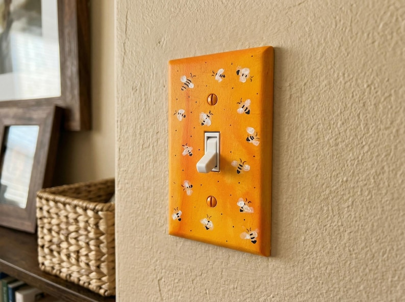 Puede incluir: Una placa de interruptor de luz rectangular con un fondo degradado de color naranja y amarillo vibrante, decorada con un patr&oacute;n repetitivo de peque&ntilde;as abejas pintadas a mano en blanco y negro. La placa se fija con dos tornillos naranjas a juego.