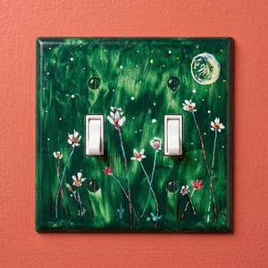 May include: A double light switch plate featuring a hand-painted dark green background with white and red wildflowers and a crescent moon. The rectangular cover has two white toggle switches and four matching green screws securing it to the wall.