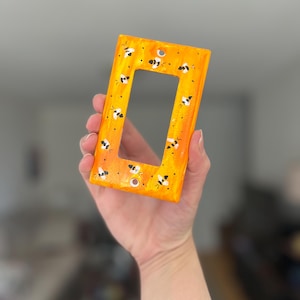 Puede incluir: Una placa rectangular para interruptor de luz con un fondo jaspeado en tonos naranja y amarillo vibrantes. La superficie est&aacute; decorada con un patr&oacute;n repetitivo de peque&ntilde;as abejas pintadas a mano en blanco y negro por toda la parte frontal.