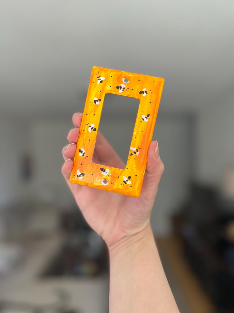 Puede incluir: Una placa rectangular para interruptor de luz con un fondo jaspeado en tonos naranja y amarillo vibrantes. La superficie est&aacute; decorada con un patr&oacute;n repetitivo de peque&ntilde;as abejas pintadas a mano en blanco y negro por toda la parte frontal.