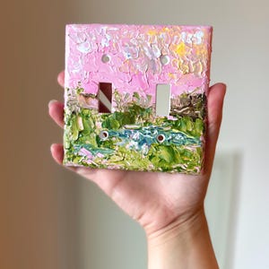 May include: A double light switch cover plate featuring a thick, textured impasto oil painting of a landscape. The design displays a vibrant pink sky above a green field with a small blue pond, applied with visible brushstrokes and paint layers.
