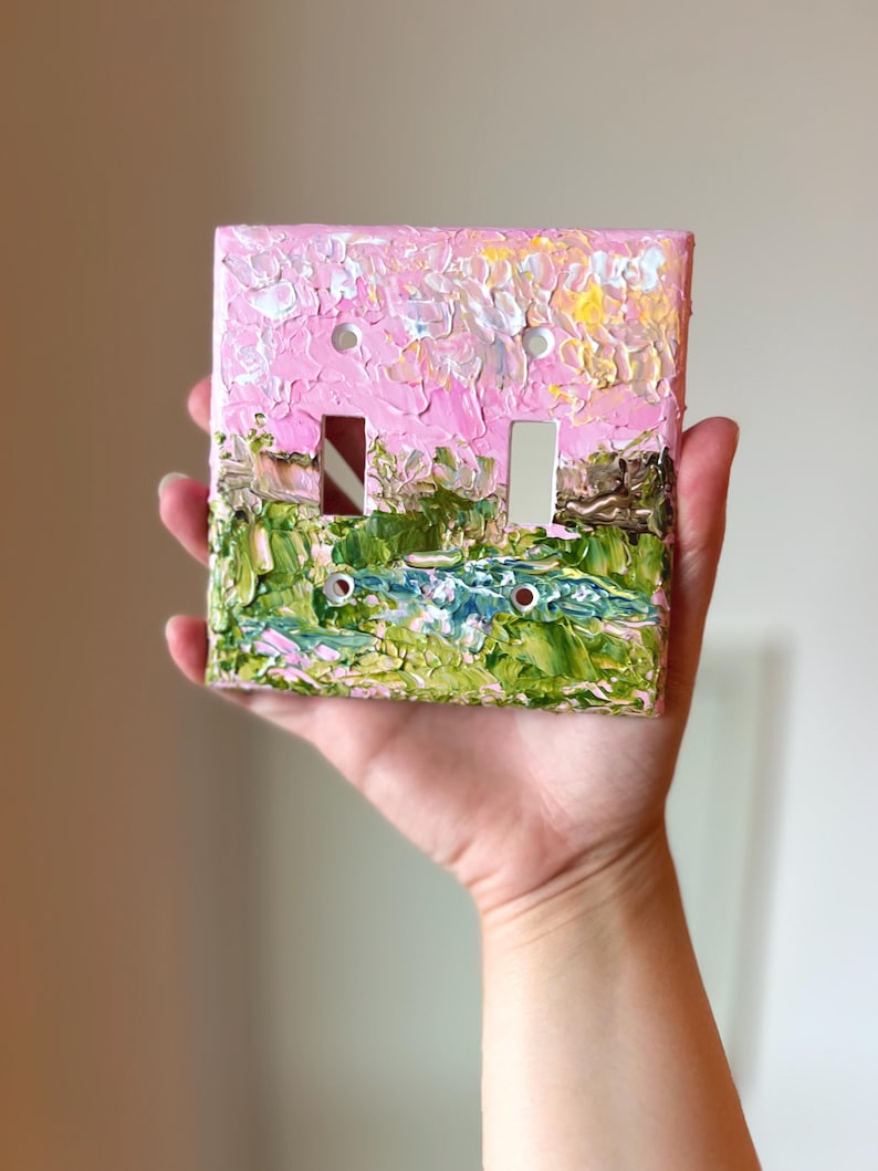 May include: A double light switch cover plate featuring a thick, textured impasto oil painting of a landscape. The design displays a vibrant pink sky above a green field with a small blue pond, applied with visible brushstrokes and paint layers.