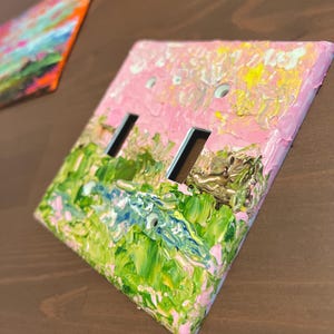 May include: A rectangular light switch cover plate featuring a thick, textured impasto oil painting application. The surface displays a vibrant landscape scene with blended strokes of pastel pink, bright yellow, forest green, and deep blue paint.