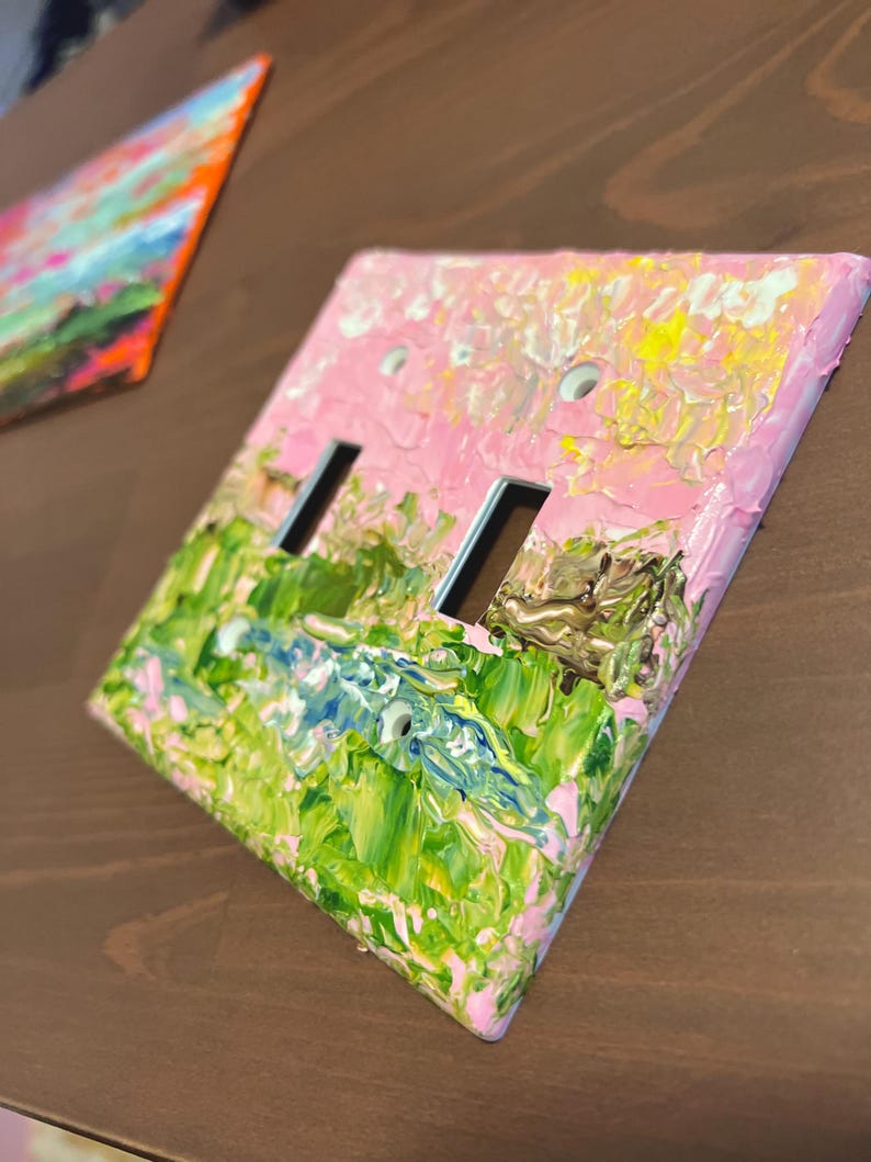 May include: A rectangular light switch cover plate featuring a thick, textured impasto oil painting application. The surface displays a vibrant landscape scene with blended strokes of pastel pink, bright yellow, forest green, and deep blue paint.