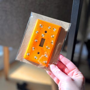 Puede incluir: Una placa de cubierta para interruptor de luz rectangular pintada en un color naranja vibrante, con un patr&oacute;n repetitivo de peque&ntilde;as abejas blancas y negras pintadas a mano sobre la superficie. La placa tiene dos orificios para tornillos y una abertura central.