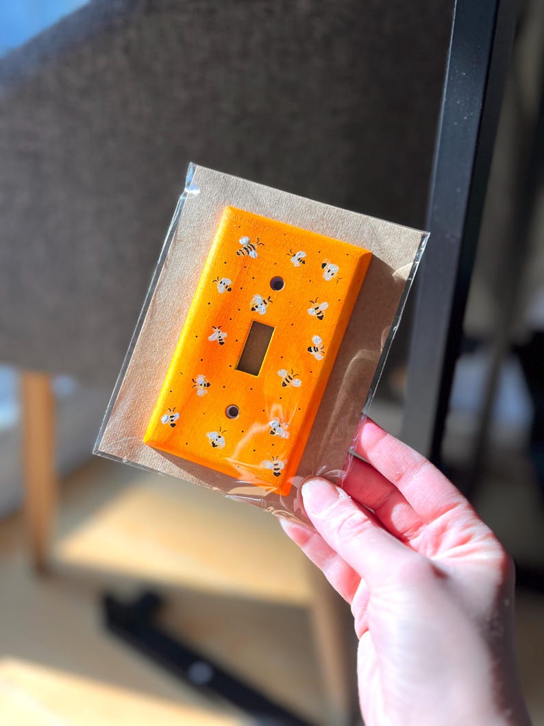 Puede incluir: Una placa de cubierta para interruptor de luz rectangular pintada en un color naranja vibrante, con un patr&oacute;n repetitivo de peque&ntilde;as abejas blancas y negras pintadas a mano sobre la superficie. La placa tiene dos orificios para tornillos y una abertura central.