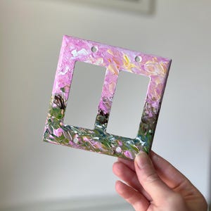 May include: A double light switch wall plate featuring a thick, textured impasto paint application. The surface displays a blend of soft pink, white, yellow, and deep green brushstrokes, creating a layered, abstract floral-like appearance.