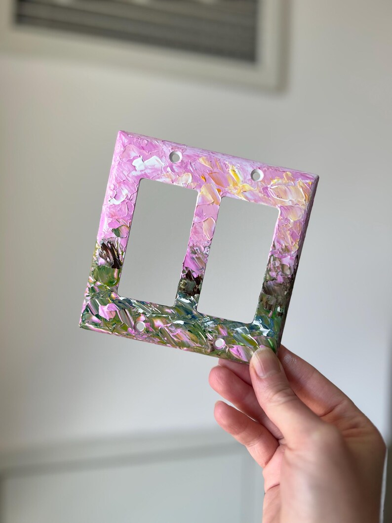 May include: A double light switch wall plate featuring a thick, textured impasto paint application. The surface displays a blend of soft pink, white, yellow, and deep green brushstrokes, creating a layered, abstract floral-like appearance.