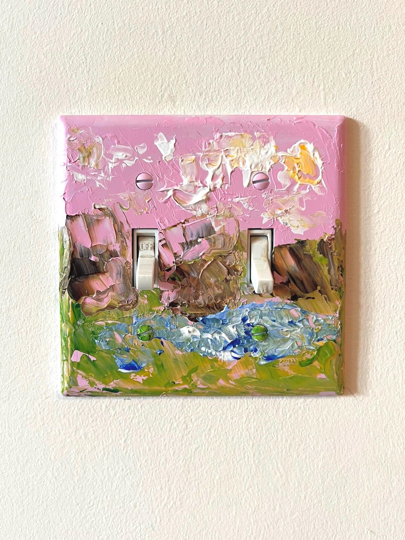 May include: A double light switch wall plate featuring a thick, textured impasto oil painting. The artwork displays a landscape with a pink sky, brown rock formations, and a blue and green grassy foreground. Two white toggle switches are centered.