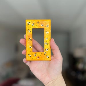 Puede incluir: Una placa rectangular para interruptor de luz con un fondo moteado en amarillo y naranja vibrante. La superficie est&aacute; decorada con un patr&oacute;n repetitivo de peque&ntilde;as abejas pintadas a mano en blanco y negro, dispersas por toda la cara frontal.