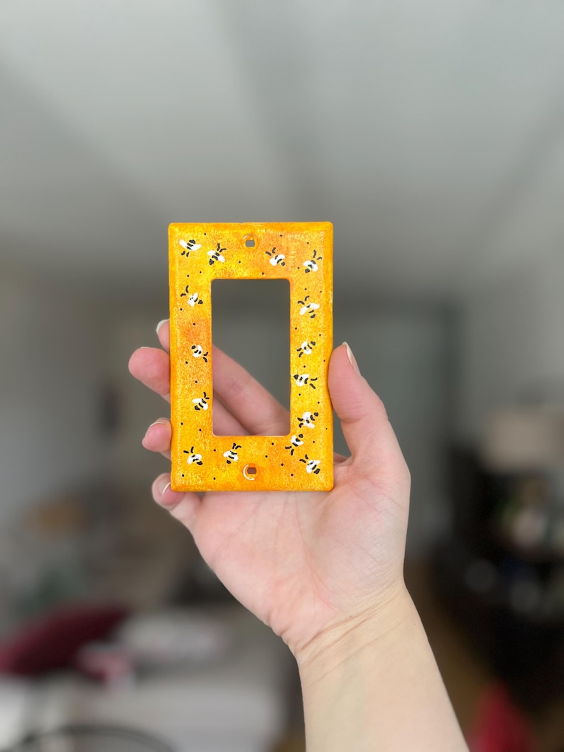 Puede incluir: Una placa rectangular para interruptor de luz con un fondo moteado en amarillo y naranja vibrante. La superficie est&aacute; decorada con un patr&oacute;n repetitivo de peque&ntilde;as abejas pintadas a mano en blanco y negro, dispersas por toda la cara frontal.