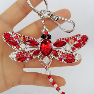 Op de afbeelding: Een sleutelhanger in de vorm van een libelle met een zilverkleurig metalen frame en rode en iriserende kristallen. De vleugels van de libelle zijn versierd met rode kristallen en het lichaam heeft een grote rode kristal. De sleutelhanger bevat een karabijnhaak en een sleutelring.