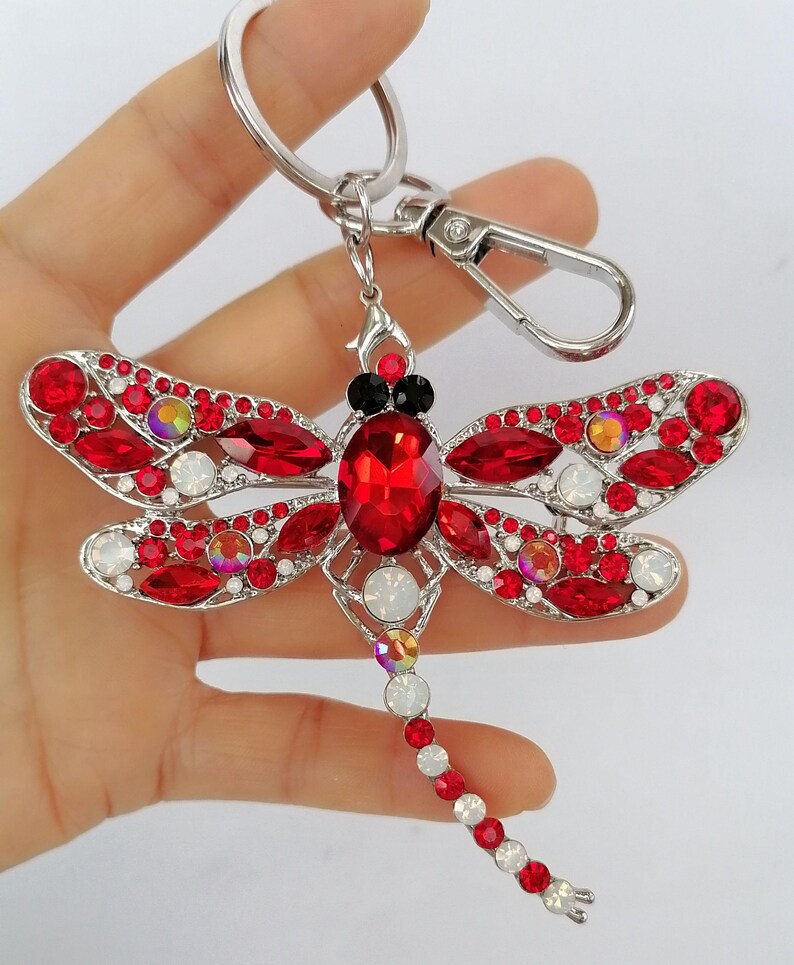 Op de afbeelding: Een sleutelhanger in de vorm van een libelle met een zilverkleurig metalen frame en rode en iriserende kristallen. De vleugels van de libelle zijn versierd met rode kristallen en het lichaam heeft een grote rode kristal. De sleutelhanger bevat een karabijnhaak en een sleutelring.