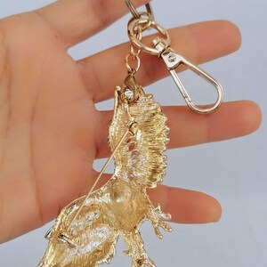 May include: Gold-toned metal keychain featuring a detailed, ornate bird brooch. The bird has textured feathers and is attached to a gold chain, a clasp, and a keyring. The brooch has a pin on the back.