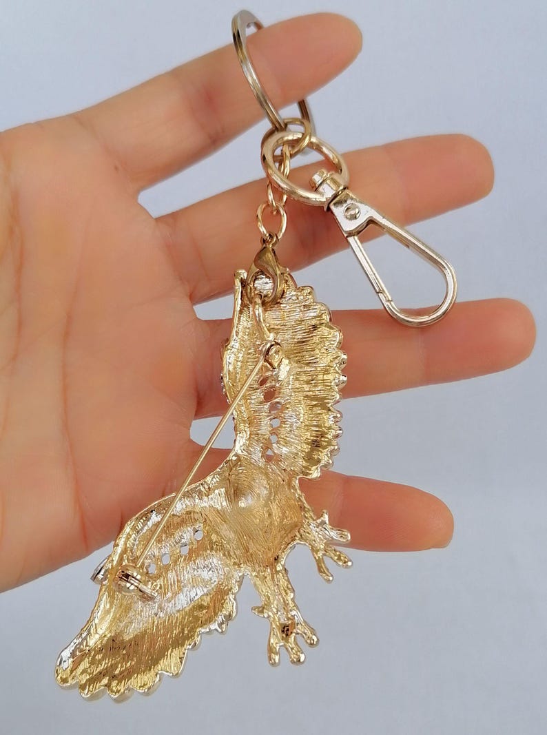 May include: Gold-toned metal keychain featuring a detailed, ornate bird brooch. The bird has textured feathers and is attached to a gold chain, a clasp, and a keyring. The brooch has a pin on the back.