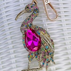 May include: A gold-toned metal keychain featuring a bejeweled bird design. The bird has a large pink gemstone on its chest and is adorned with colorful rhinestones. The keychain includes a clasp and a ring.