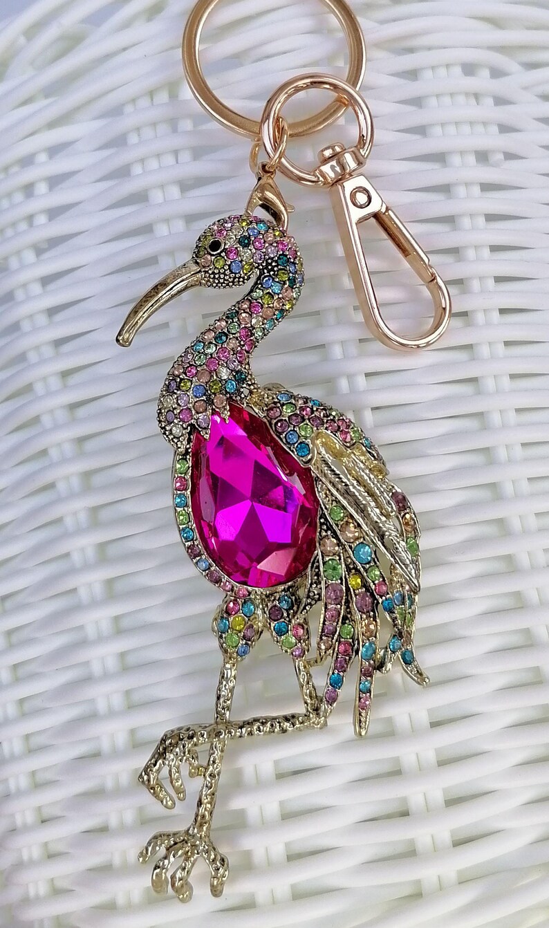 May include: A gold-toned metal keychain featuring a bejeweled bird design. The bird has a large pink gemstone on its chest and is adorned with colorful rhinestones. The keychain includes a clasp and a ring.
