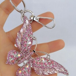 May include: A pink butterfly keychain with a silver-tone metal finish. The butterfly is encrusted with pink rhinestones and has a lobster clasp and key ring for attachment. The wings are detailed with various shapes of pink crystals.