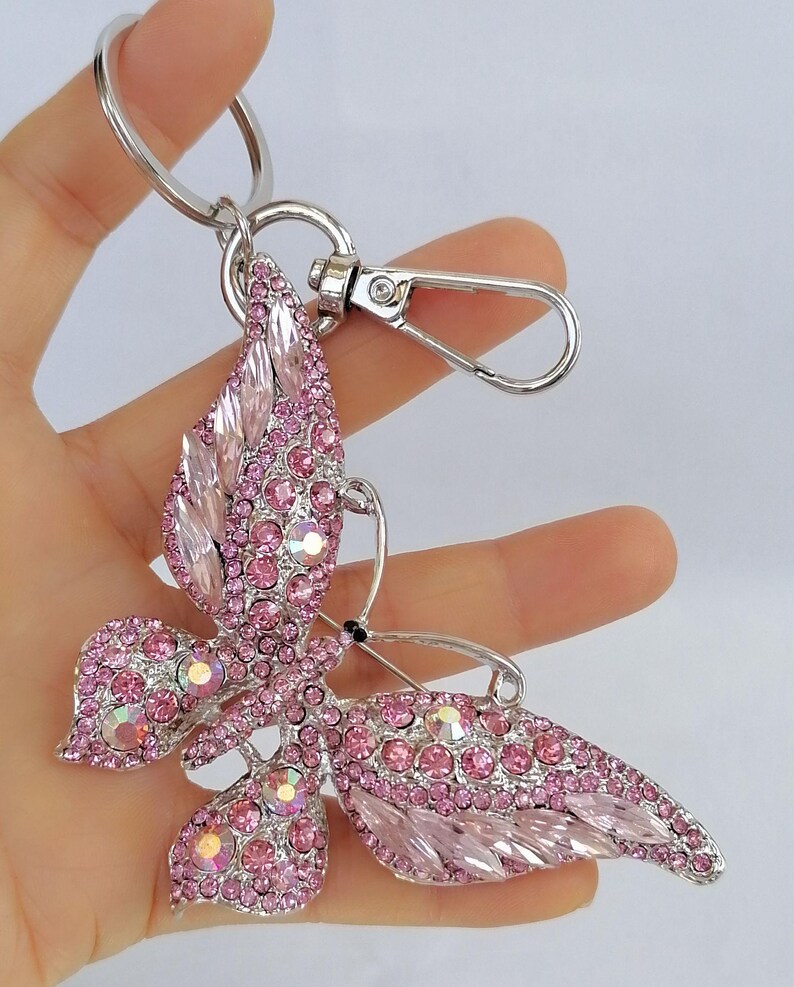 May include: A pink butterfly keychain with a silver-tone metal finish. The butterfly is encrusted with pink rhinestones and has a lobster clasp and key ring for attachment. The wings are detailed with various shapes of pink crystals.