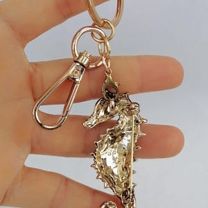 K&ouml;nnte beinhalten: Goldfarbener Seepferdchen-Schl&uuml;sselanh&auml;nger mit Karabinerverschluss und Schl&uuml;sselring. Der Seepferdchen-Anh&auml;nger hat ein detailliertes, strukturiertes Design. Der Schl&uuml;sselanh&auml;nger wird in einer Hand vor hellem Hintergrund gehalten.