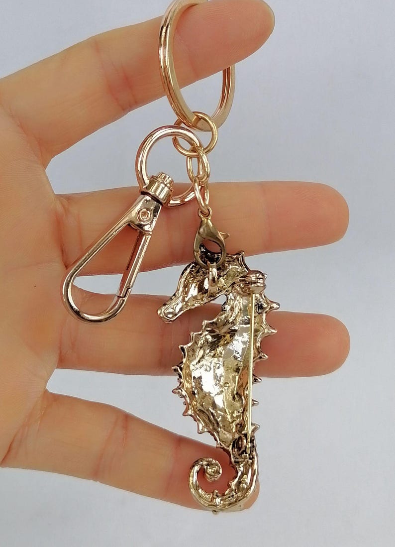 K&ouml;nnte beinhalten: Goldfarbener Seepferdchen-Schl&uuml;sselanh&auml;nger mit Karabinerverschluss und Schl&uuml;sselring. Der Seepferdchen-Anh&auml;nger hat ein detailliertes, strukturiertes Design. Der Schl&uuml;sselanh&auml;nger wird in einer Hand vor hellem Hintergrund gehalten.