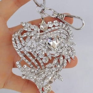 May include: Silver-toned heart-shaped keychain adorned with numerous clear crystals. The design features a central large crystal and smaller crystals arranged in a floral pattern. Includes a keyring and a lobster clasp.