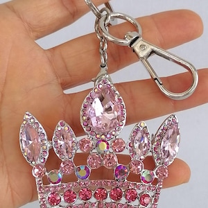 May include: A silver-toned keychain featuring a pink jeweled crown pendant. The crown is adorned with various sizes of pink rhinestones and a large teardrop-shaped pink gem at the top. A silver clasp and key ring are attached.