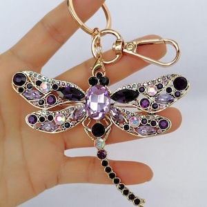 Op de afbeelding: Een goudkleurige sleutelhanger in de vorm van een libelle, versierd met paarse en iriserende kristallen. De libelle heeft een groot ovaal kristallen lichaam, kristallen vleugels en een reeks kleinere kristallen langs de staart. De sleutelhanger bevat een sluiting en een ring.