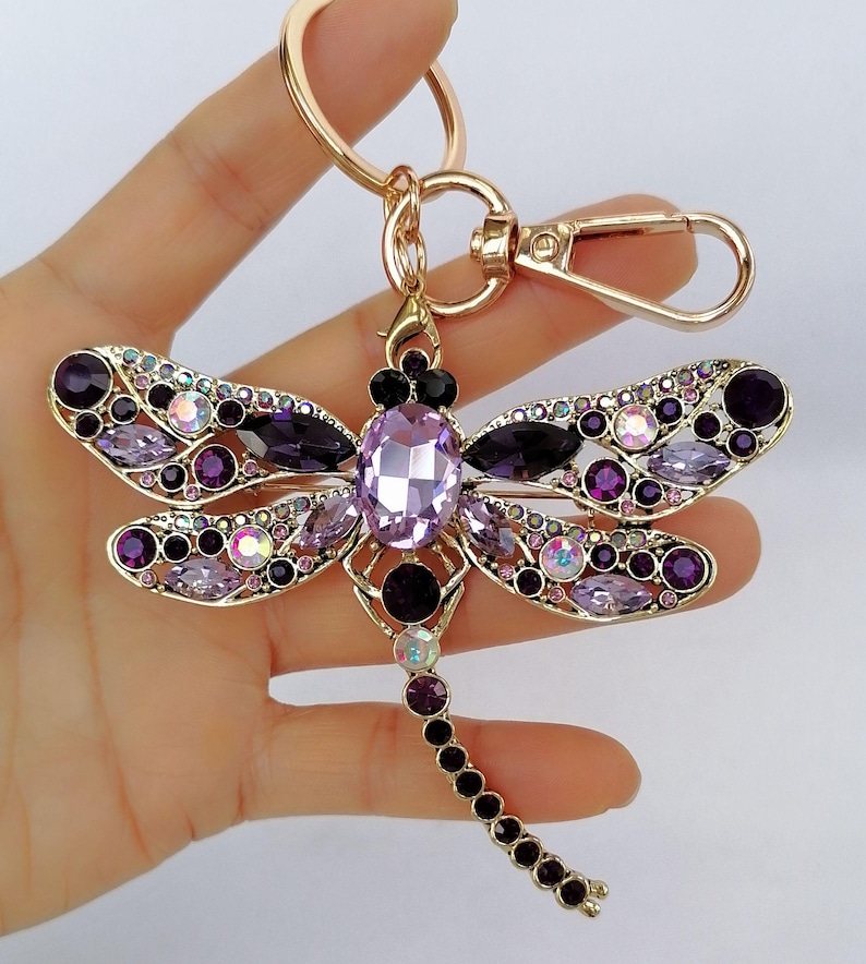 Op de afbeelding: Een goudkleurige sleutelhanger in de vorm van een libelle, versierd met paarse en iriserende kristallen. De libelle heeft een groot ovaal kristallen lichaam, kristallen vleugels en een reeks kleinere kristallen langs de staart. De sleutelhanger bevat een sluiting en een ring.