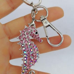 K&ouml;nnte beinhalten: Ein silberner Schl&uuml;sselanh&auml;nger mit einem Anh&auml;nger in Form eines Seepferdchens, besetzt mit rosa und irisierenden Strasssteinen. Das Seepferdchen hat ein schwarzes Auge und ist an einem Spaltring und einem Karabiner befestigt.