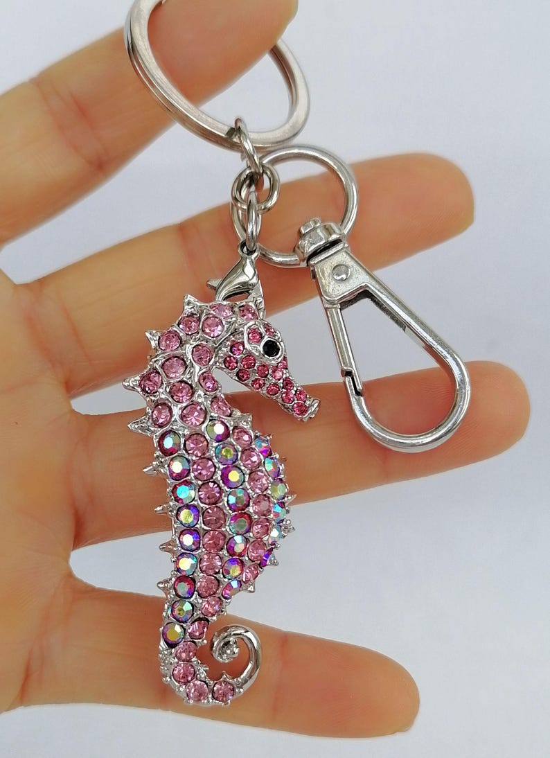 K&ouml;nnte beinhalten: Ein silberner Schl&uuml;sselanh&auml;nger mit einem Anh&auml;nger in Form eines Seepferdchens, besetzt mit rosa und irisierenden Strasssteinen. Das Seepferdchen hat ein schwarzes Auge und ist an einem Spaltring und einem Karabiner befestigt.