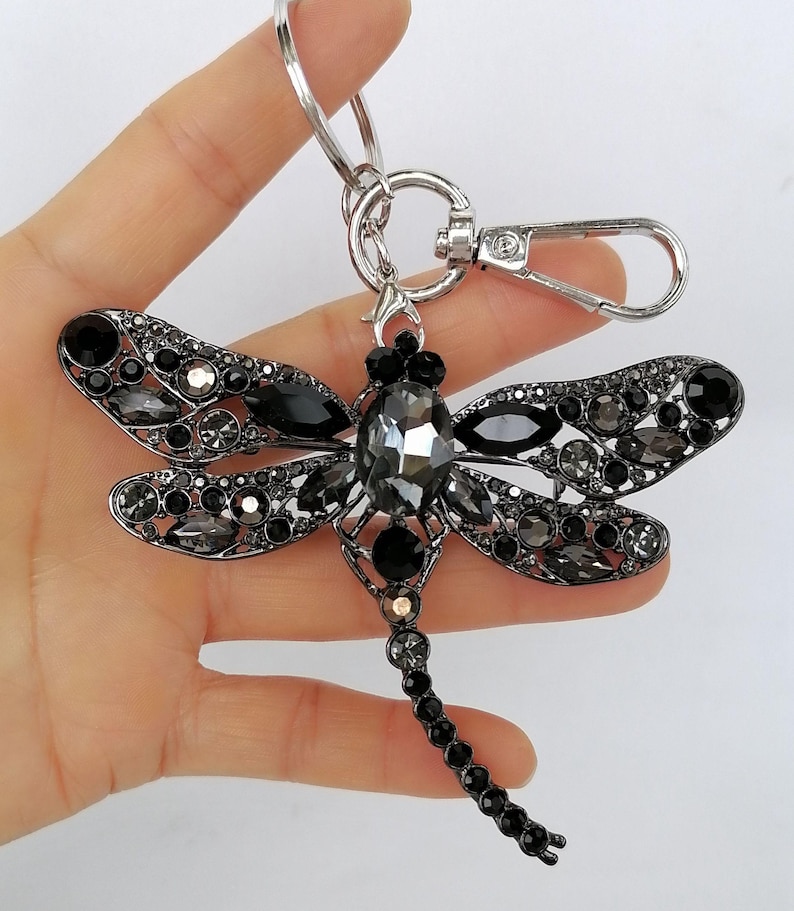 Op de afbeelding: Een sleutelhanger in de vorm van een libelle met een zilverkleurige metalen afwerking. De libelle is versierd met zwarte en grijze kristallen. De sleutelhanger heeft een karabijnhaak en een sleutelring.