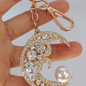 May include: Gold-toned keychain featuring a crescent moon design adorned with clear crystals and a pearl. The keychain has a lobster clasp and a ring for attaching keys. A decorative accessory.