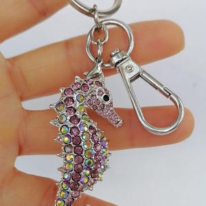 K&ouml;nnte beinhalten: Ein silberfarbener Schl&uuml;sselanh&auml;nger in Form eines Seepferdchens mit Karabinerverschluss und Schl&uuml;sselring. Das Seepferdchen ist mit rosa und irisierenden Kristallen besetzt und hat ein schwarzes Auge. Der Schl&uuml;sselanh&auml;nger wird vor wei&szlig;em Hintergrund gehalten.