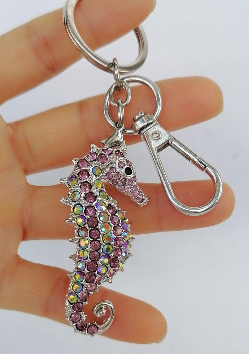 K&ouml;nnte beinhalten: Ein silberfarbener Schl&uuml;sselanh&auml;nger in Form eines Seepferdchens mit Karabinerverschluss und Schl&uuml;sselring. Das Seepferdchen ist mit rosa und irisierenden Kristallen besetzt und hat ein schwarzes Auge. Der Schl&uuml;sselanh&auml;nger wird vor wei&szlig;em Hintergrund gehalten.