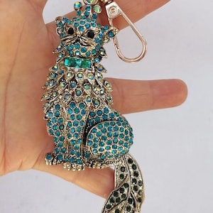 May include: A jeweled cat keychain in shades of teal and gold. The cat is adorned with numerous teal-colored rhinestones and has a green rectangular gem on its collar. The tail is a stylized leaf design with green gems.