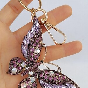 May include: A butterfly-shaped keychain with a gold-tone ring and clasp. The butterfly is adorned with purple and clear crystals, creating a sparkling effect. The keychain is a decorative accessory.