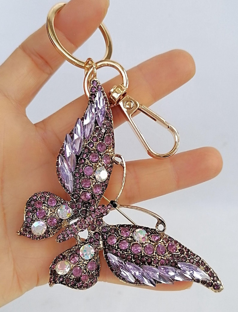 May include: A butterfly-shaped keychain with a gold-tone ring and clasp. The butterfly is adorned with purple and clear crystals, creating a sparkling effect. The keychain is a decorative accessory.