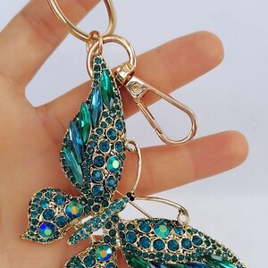 May include: A gold-toned metal keychain featuring a butterfly design. The butterfly is adorned with teal and blue crystals, creating a sparkling effect. The keychain includes a ring and a clasp for easy attachment.