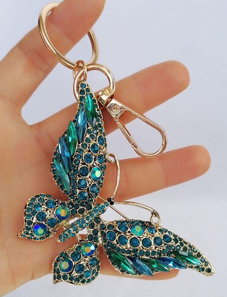 May include: A gold-toned metal keychain featuring a butterfly design. The butterfly is adorned with teal and blue crystals, creating a sparkling effect. The keychain includes a ring and a clasp for easy attachment.