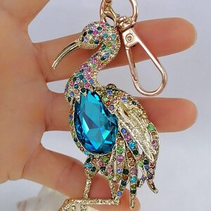 May include: A jeweled bird keychain with a gold-tone finish. The bird features a large, teardrop-shaped blue crystal and is adorned with colorful rhinestones. The keychain includes a clasp and a ring for keys.