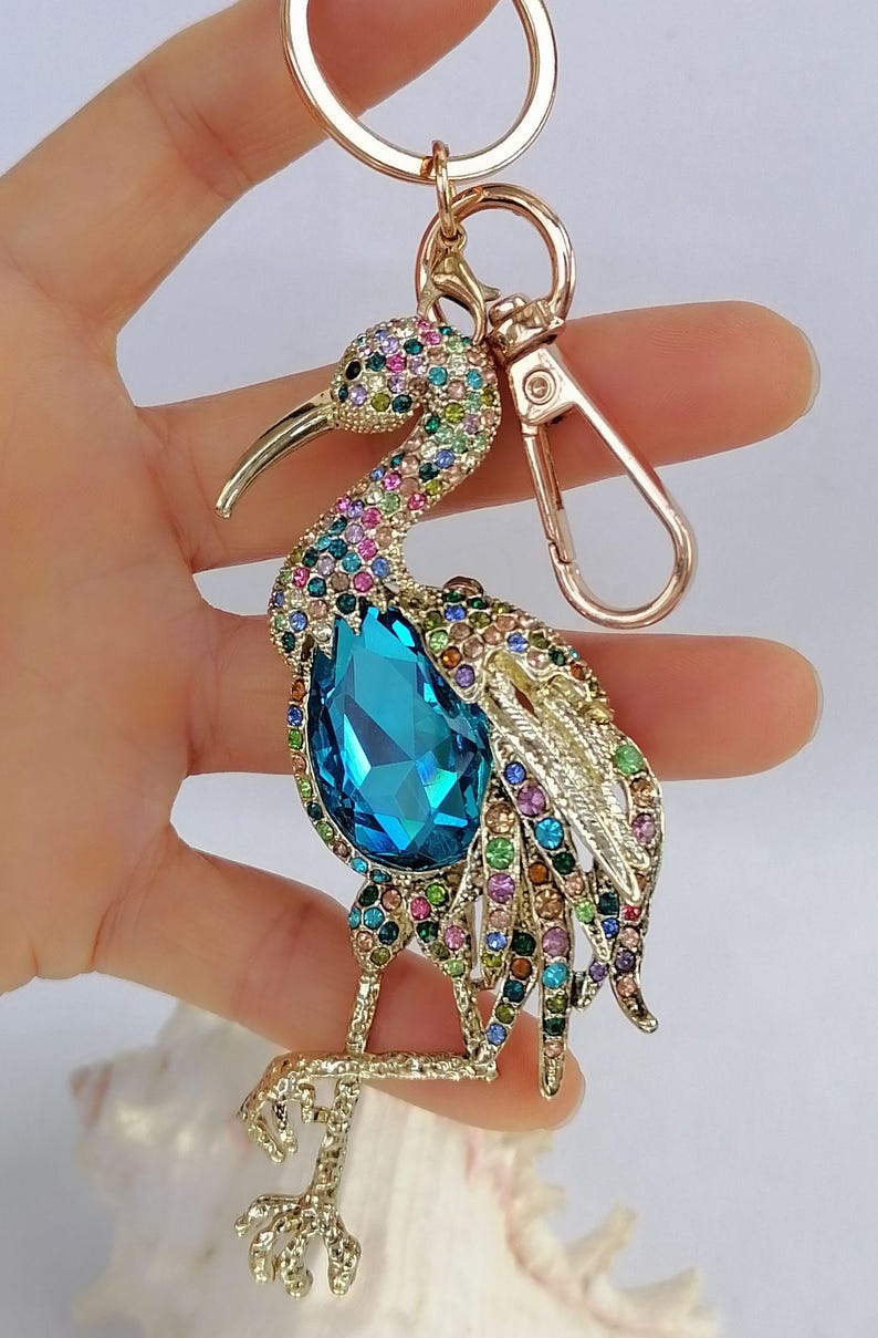 May include: A jeweled bird keychain with a gold-tone finish. The bird features a large, teardrop-shaped blue crystal and is adorned with colorful rhinestones. The keychain includes a clasp and a ring for keys.